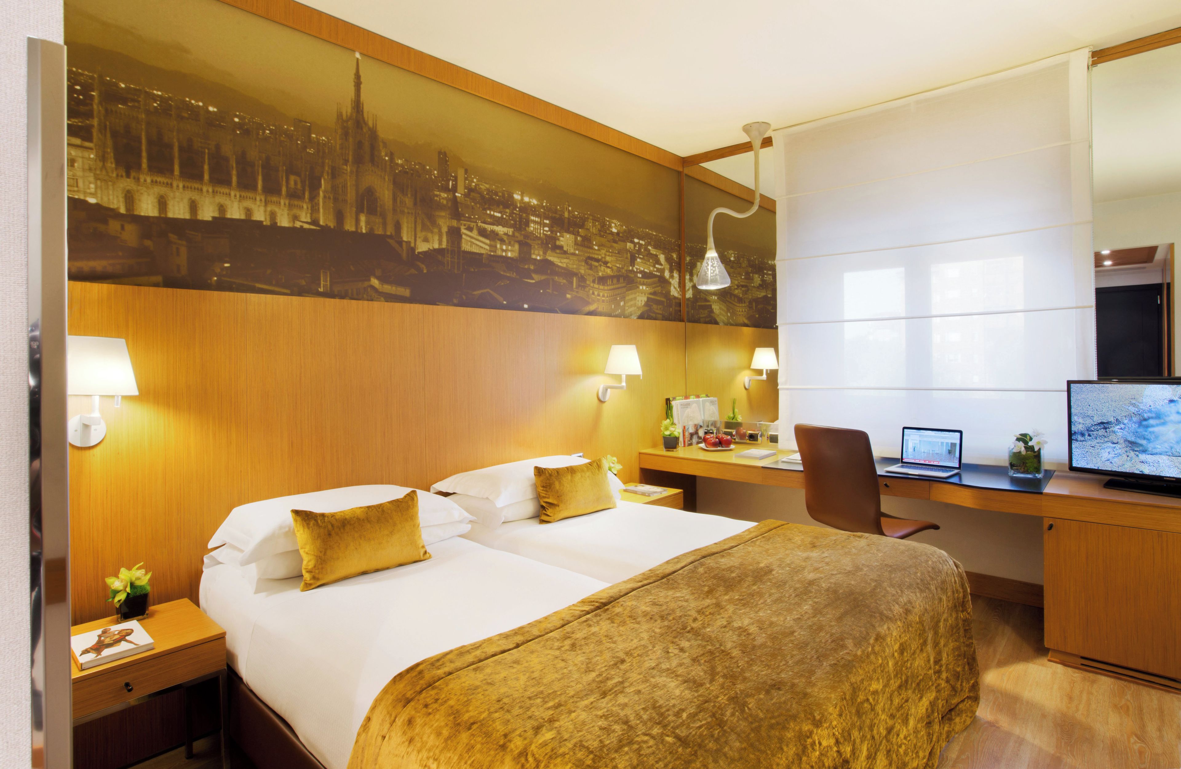 Quarto de hotel com cama, escrivaninha, computador e um grande mural com vista para a cidade.