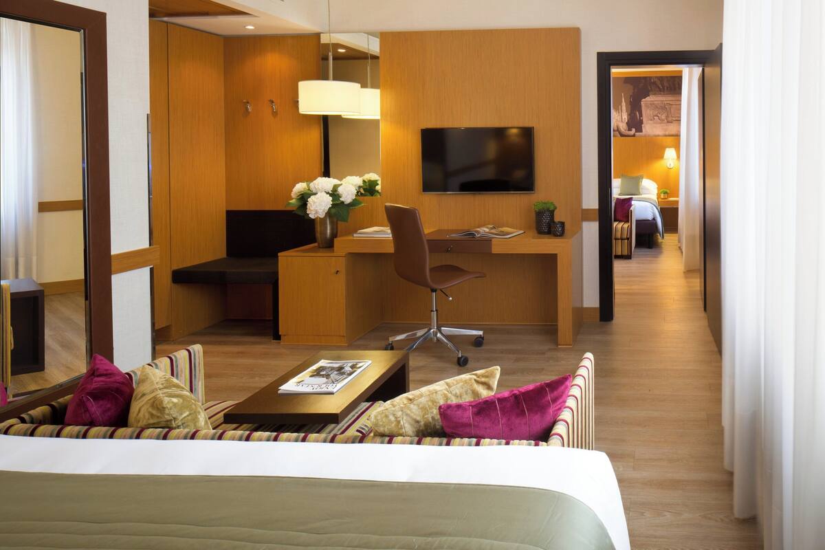 A hotel room with a bed, a desk with a chair, a TV, and a small sitting area with a sofa and cushions.