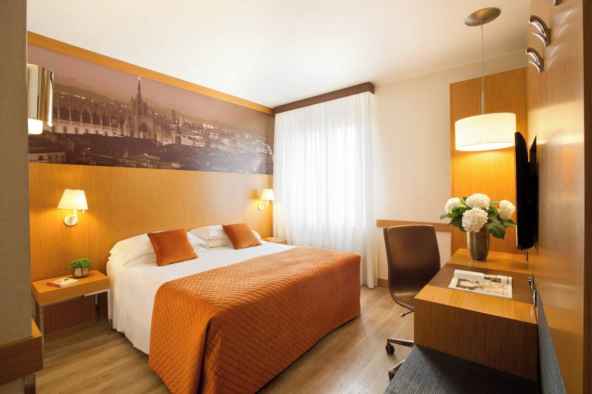 A hotel room with a large bed, two bedside tables with lamps, a desk with a chair, a vase with flowers, and a cityscape mural on the wall.