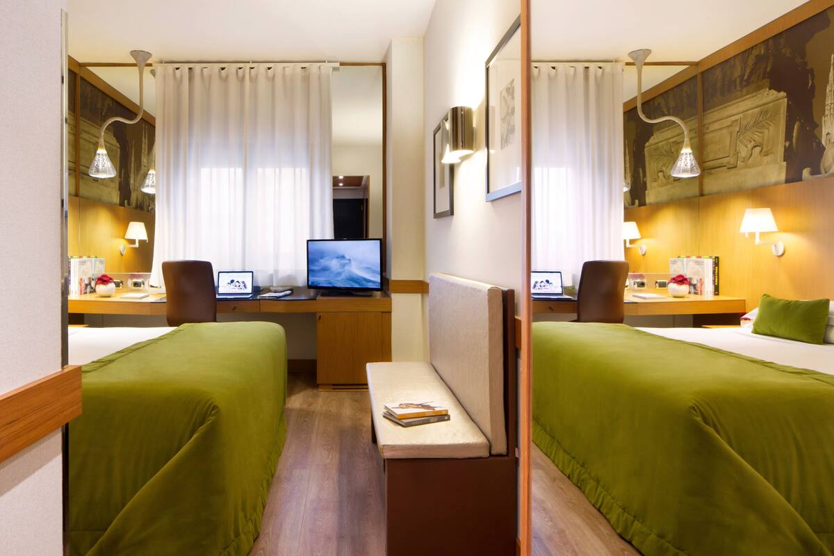 A hotel room with two beds, a desk with a computer, a chair, a small table, and a mirror.