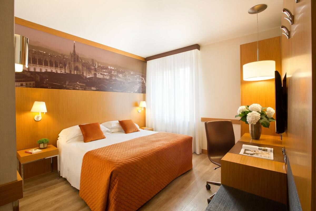 A hotel room with a bed, two bedside tables, a desk with a chair, a TV, and a cityscape mural on the wall.