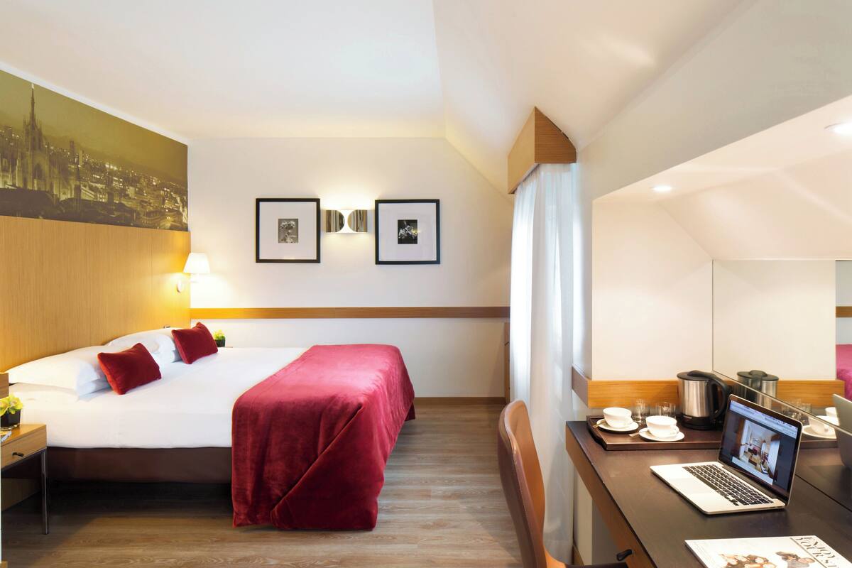 A hotel room with a bed, a desk with a laptop, a coffee maker, and framed pictures on the wall.
