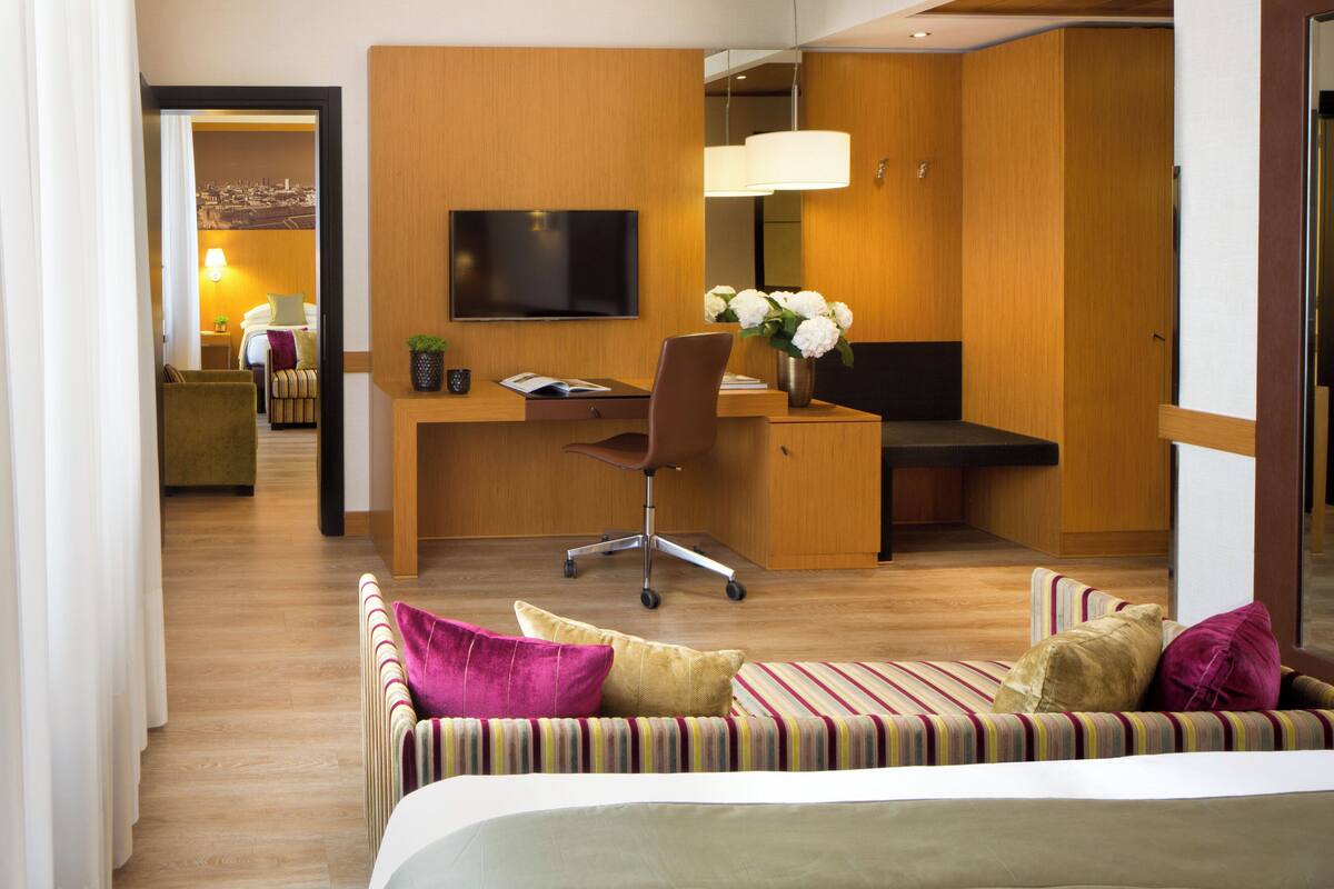 A hotel room with a bed, a desk with a chair, a television, and a view of a cityscape through an open door.