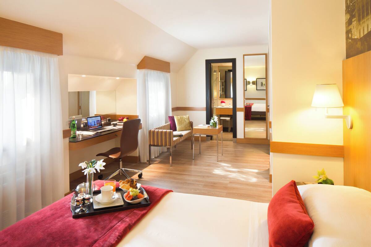 A hotel room with a bed, a desk with a laptop, a chair, a small table, and a mirror.