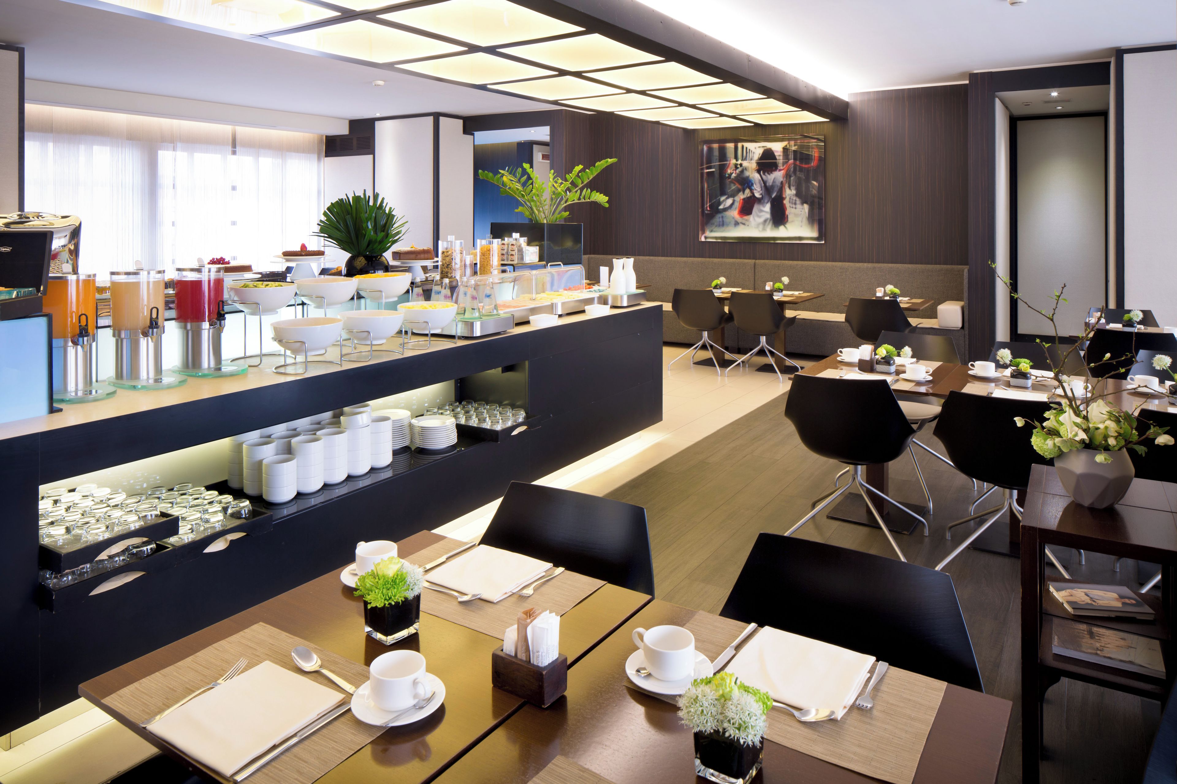 Un espace de restauration moderne dans un hôtel, avec un buffet, des tables et des chaises, et un grand écran diffusant un événement sportif.