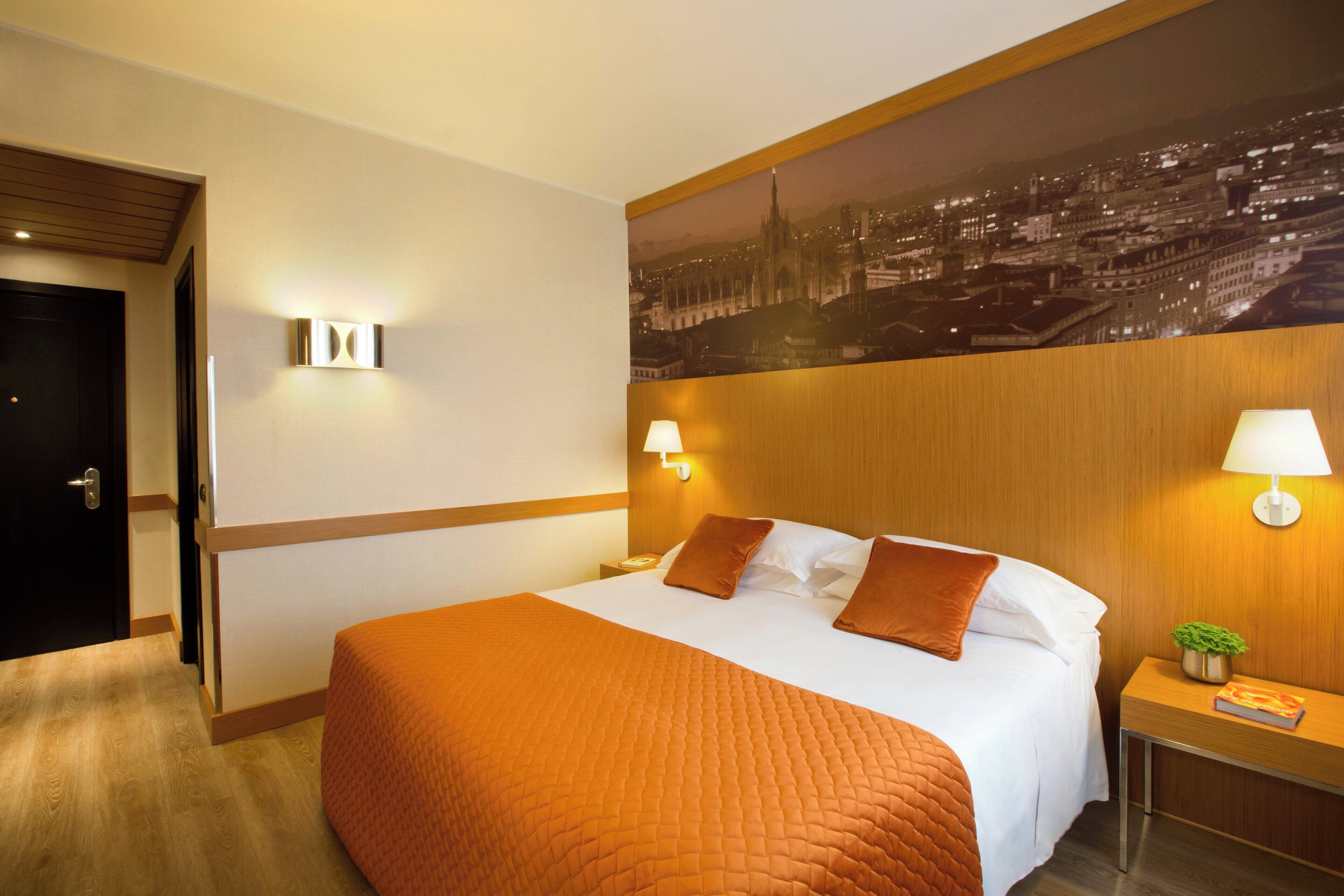Habitación de hotel con una cama grande, dos almohadas, una mesita de noche con lámpara, una pequeña mesa con una planta y un cuadro de un paisaje urbano en la pared.