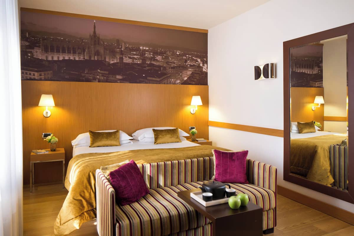 A hotel room with a large bed, a striped sofa, a small table with a tray, and a cityscape mural on the wall.
