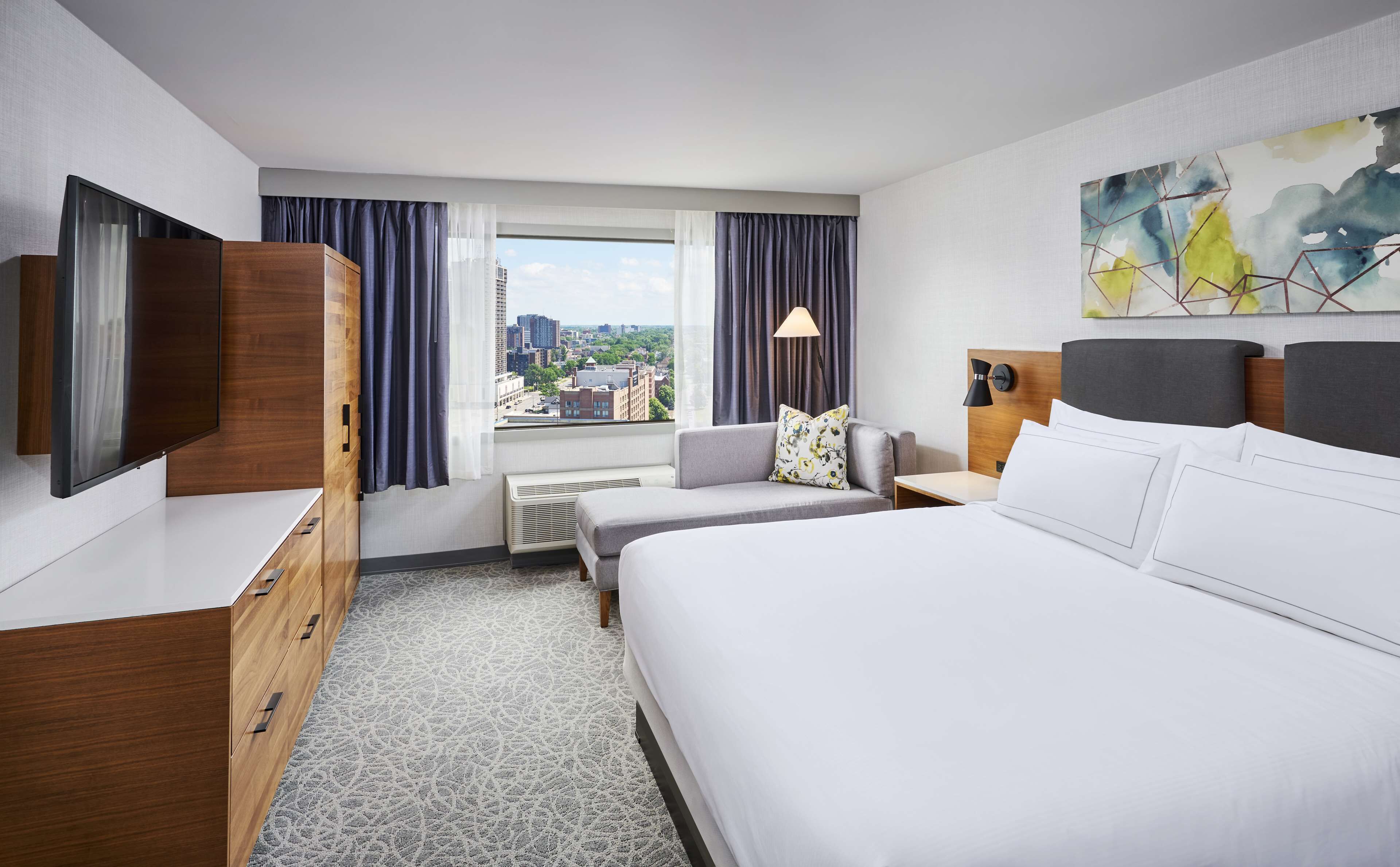 Une chambre d’hôtel avec un lit, un bureau, une chaise, une télévision et une fenêtre avec des rideaux.