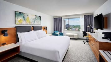 A hotel room with a large bed, a desk, a chair, a TV, and a window with a city view.
