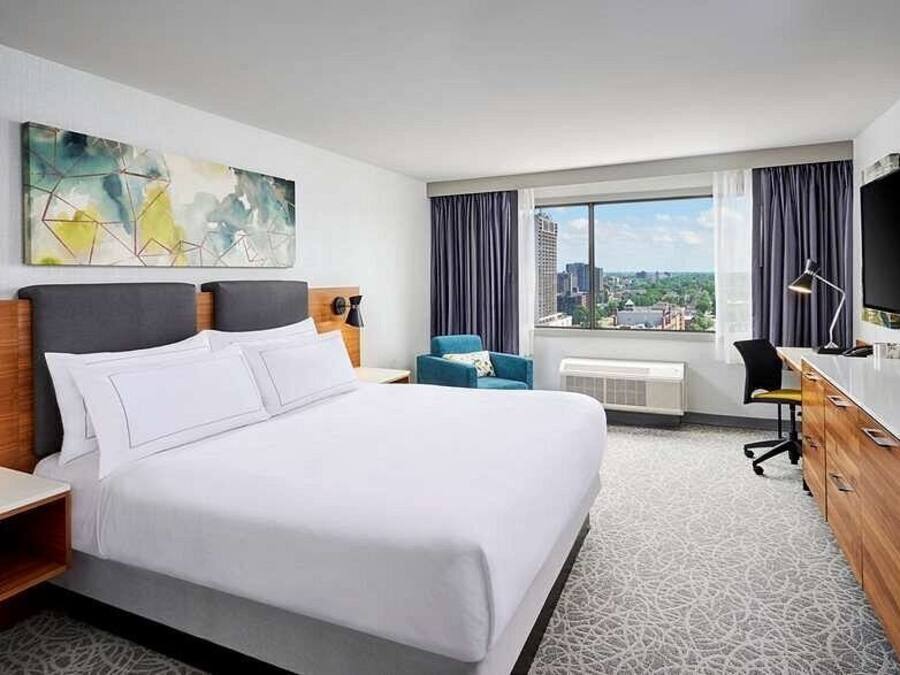 Quarto de hotel com cama grande, escrivaninha, cadeira, televisão e janela com vista para a cidade.