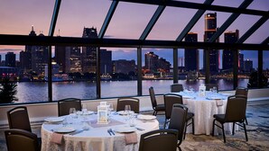 A restaurant with tables set for dining, large windows offering a city skyline view, and a river in the background.
