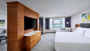 A hotel room with a large bed, a wooden wardrobe, a flat-screen TV, a desk with a chair, and a view of a cityscape through the window.