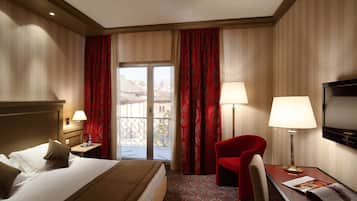Ein Hotelzimmer mit einem Bett, zwei Nachttischen mit Lampen, einem Fernseher, einem roten Sessel und einem Balkon mit roten Vorhängen.