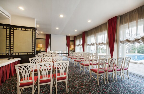 A conference room with white chairs and a projector screen, large windows with red curtains, and a pool view.