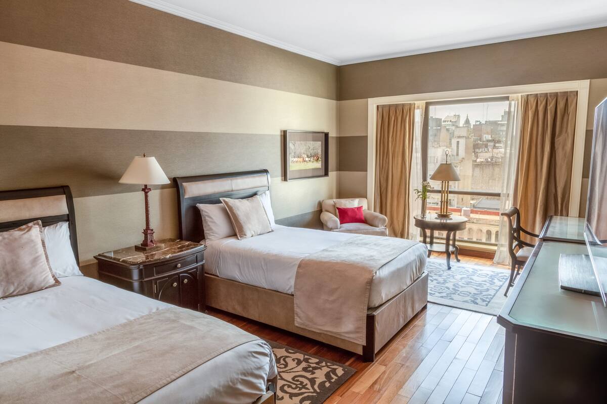 A hotel room with two beds, a desk, a chair, a small table, and a balcony with curtains.