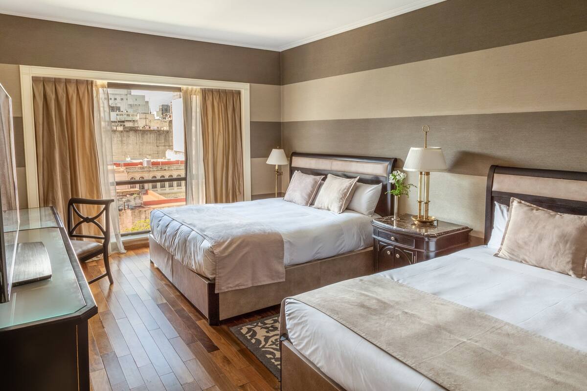 A hotel room with two beds, a desk, and a view of the cityscape through large windows.