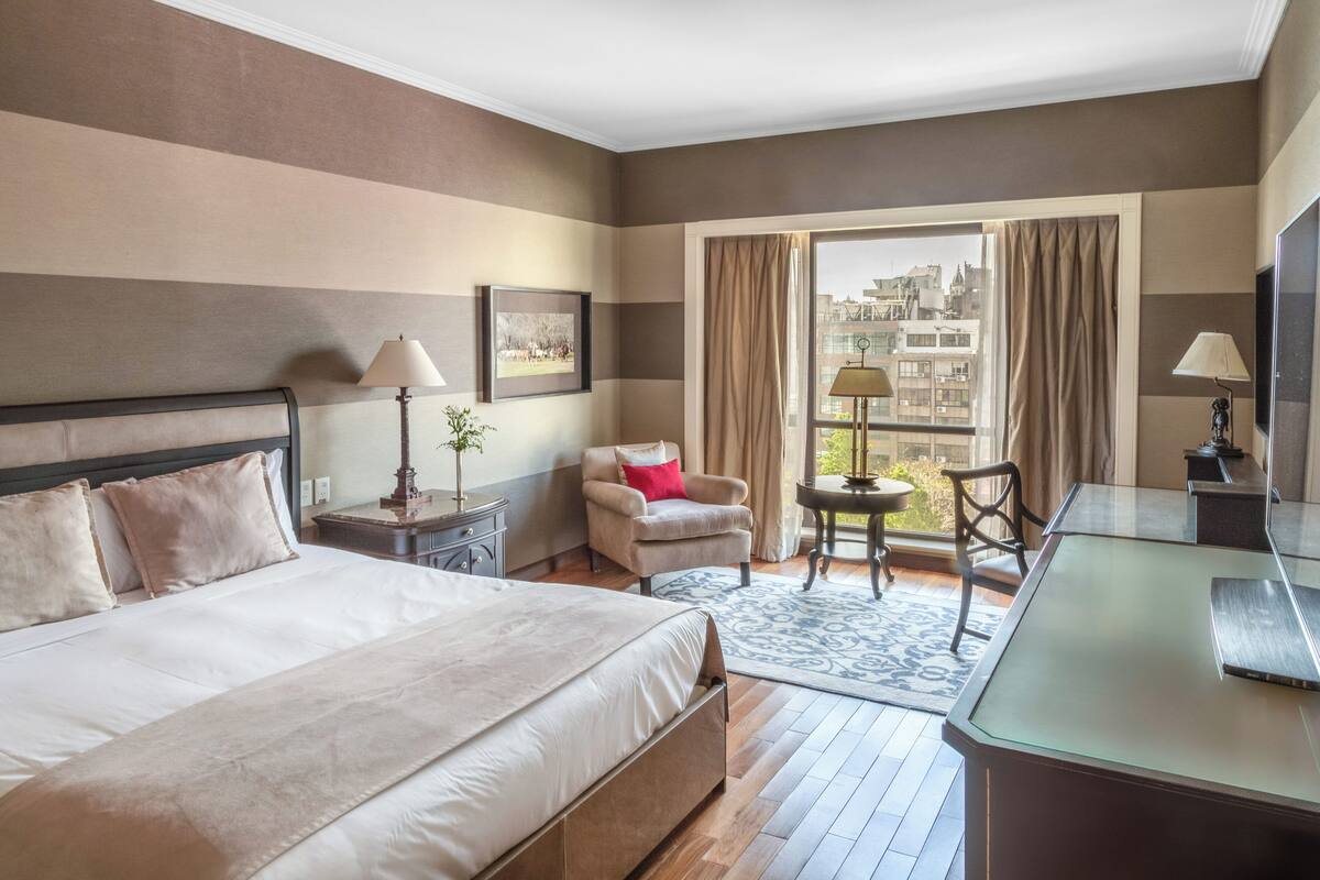 A hotel room with a large bed, a desk, a chair, a small table, a lamp, and a window with curtains.