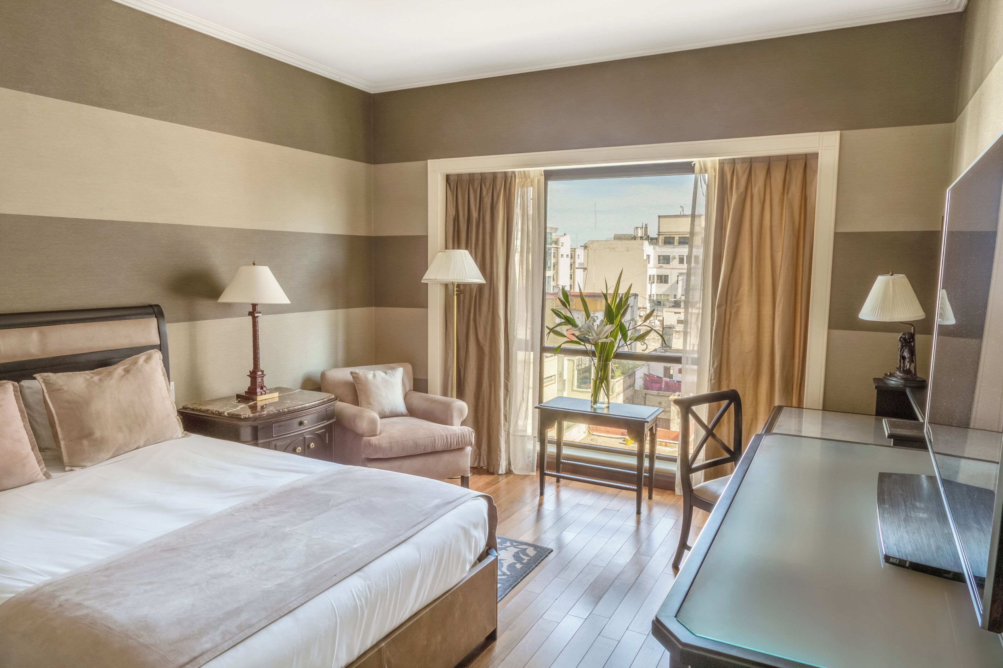 A hotel room with a bed, a desk, a chair, a small table, and a view of the city through a window.