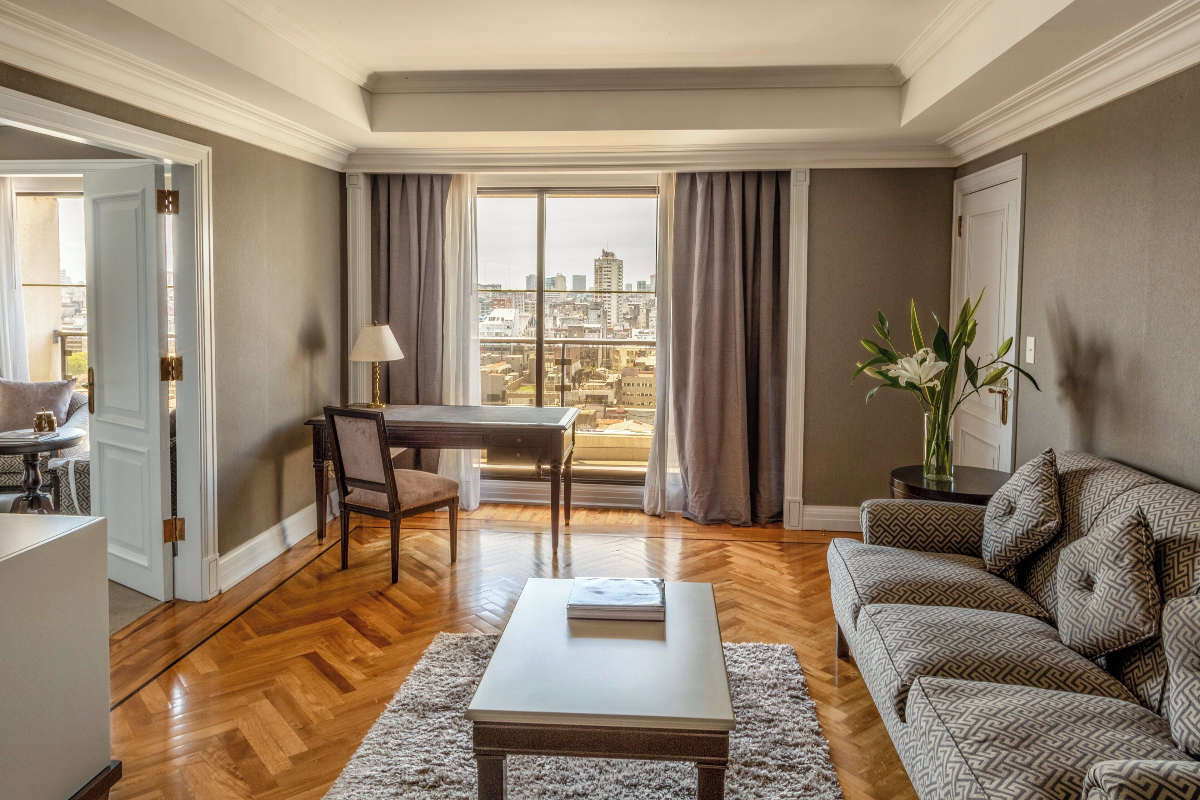 A living room with a wooden floor, a sofa, a coffee table, a dining table with chairs, and a view of a cityscape through large windows.