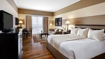 A hotel room with a large bed, a desk with a television, a chair, a nightstand with a lamp, and a window with curtains.