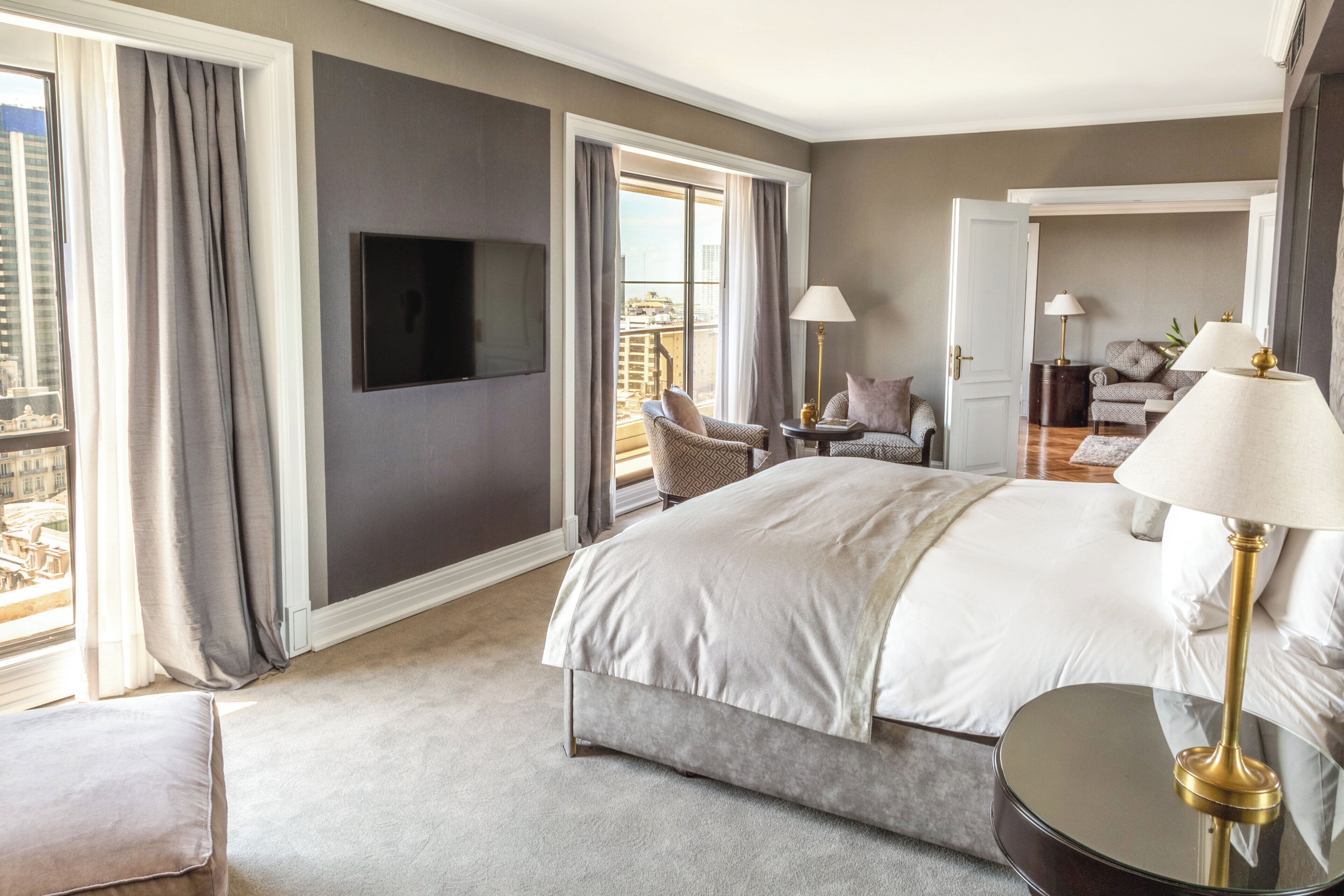 Habitación de hotel con una cama grande, televisor montado en la pared, balcón con vistas y zona de estar con una mesa y sillas.