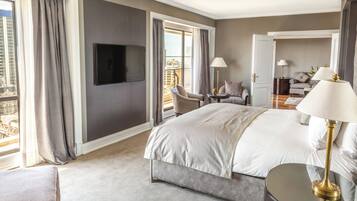 Habitación de hotel con una cama grande, televisor montado en la pared, balcón con vistas y zona de estar con una mesa y sillas.
