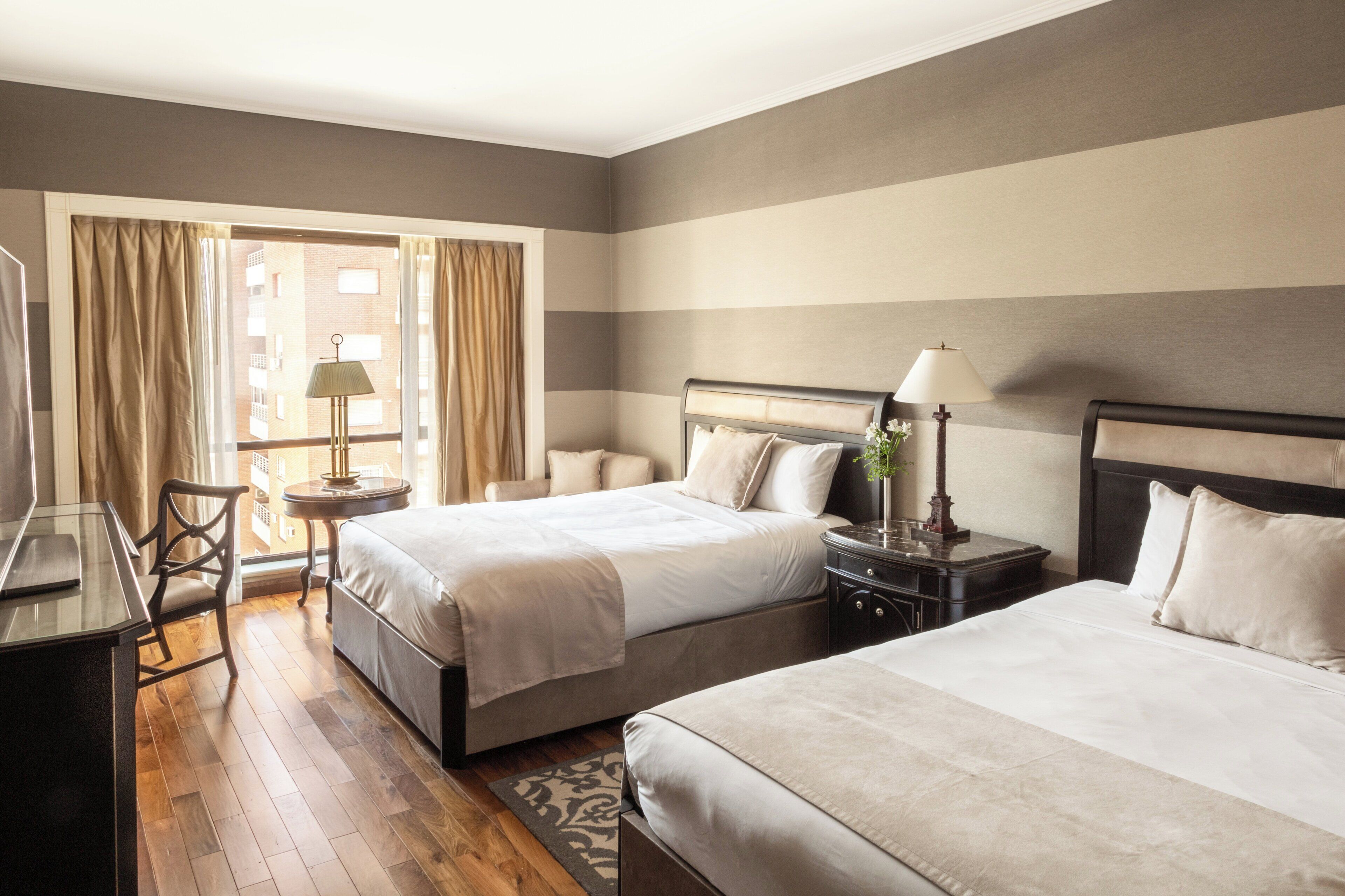 Ein Hotelzimmer mit zwei Betten, einem Schreibtisch, einem Stuhl, einer Lampe und Blick ins Freie.