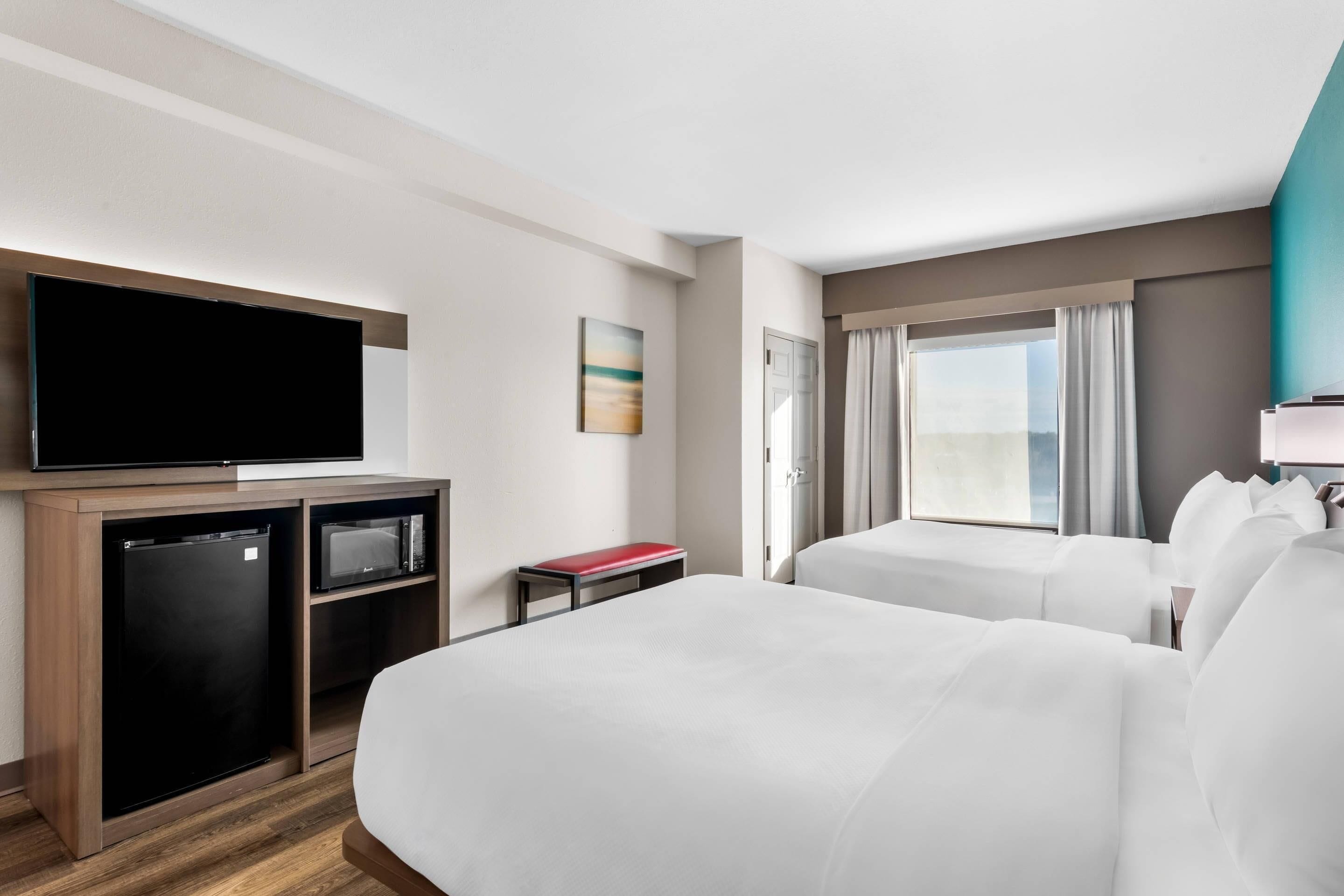 A hotel room with a bed, a TV, a mini-fridge, a microwave, a small table, and a window with curtains.