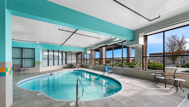 Una piscina interior con paredes de vidrio transparente, suelo de baldosas y un área de estar con sillas y mesas.