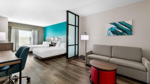 Una habitación de hotel con una cama, un escritorio, una silla, un sofá, una mesita de noche roja y un cuadro en la pared.
