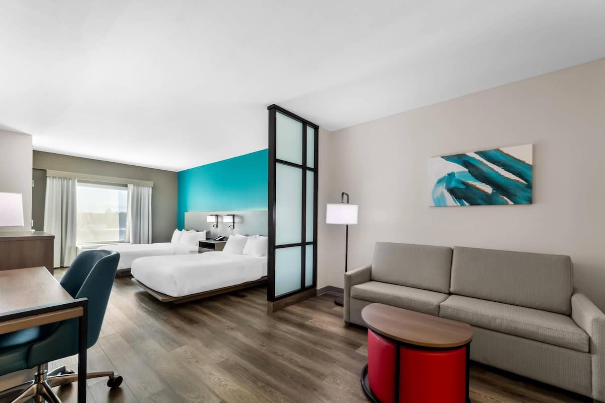 Une chambre d’hôtel avec un lit, un bureau, une chaise, un canapé, une table de chevet rouge et un tableau au mur.