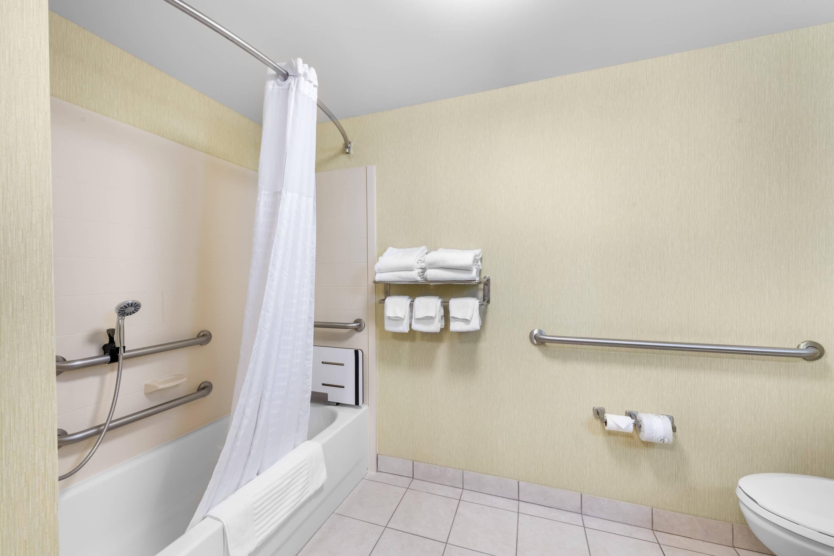 A bathroom with a bathtub, a shower with a curtain, a toilet, and handrails.