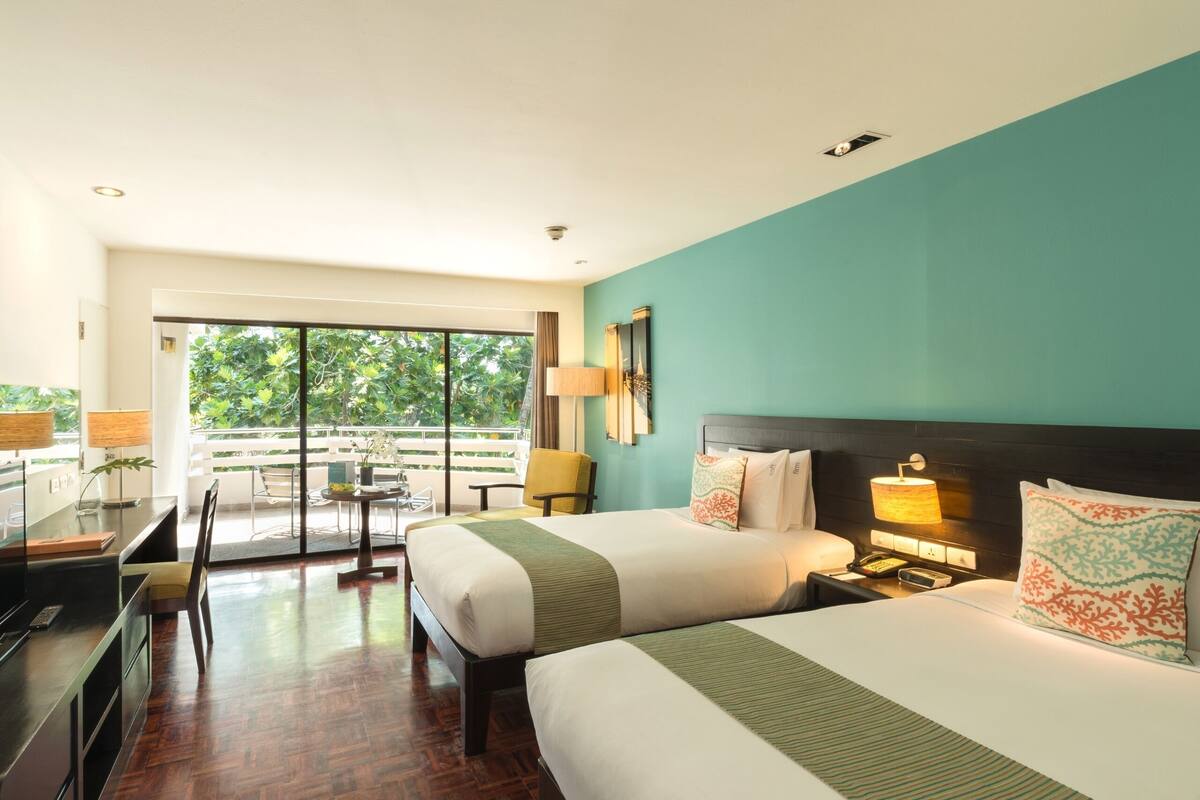 A hotel room with two beds, a desk, a chair, and a balcony with a view of greenery.