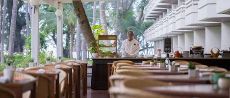 A restaurant with a chef preparing food, surrounded by tables and chairs, with a view of palm trees and a modern architectural design.