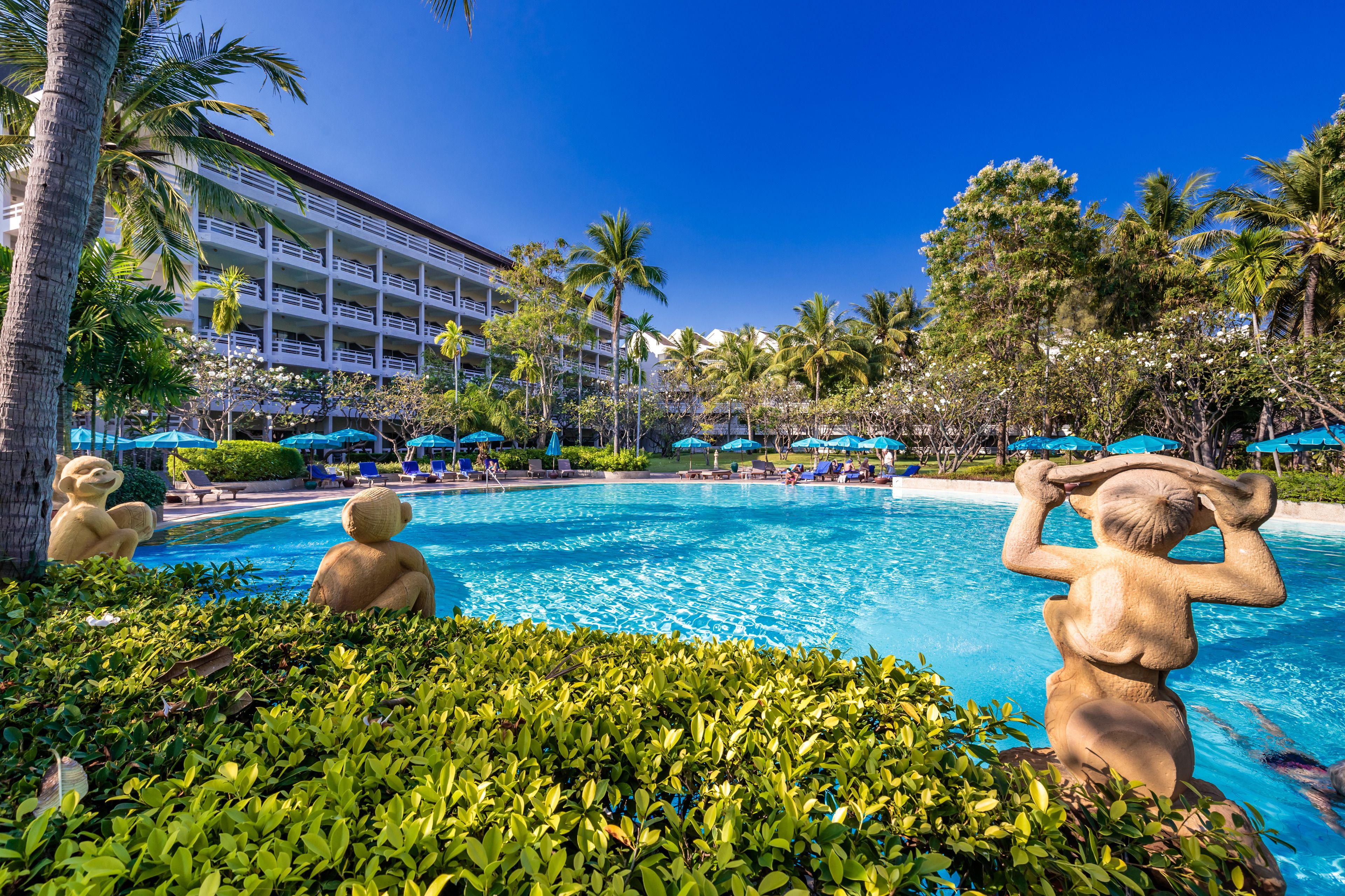 Zona de piscina de un hotel con palmeras, sillas de descanso y un gran edificio al fondo.