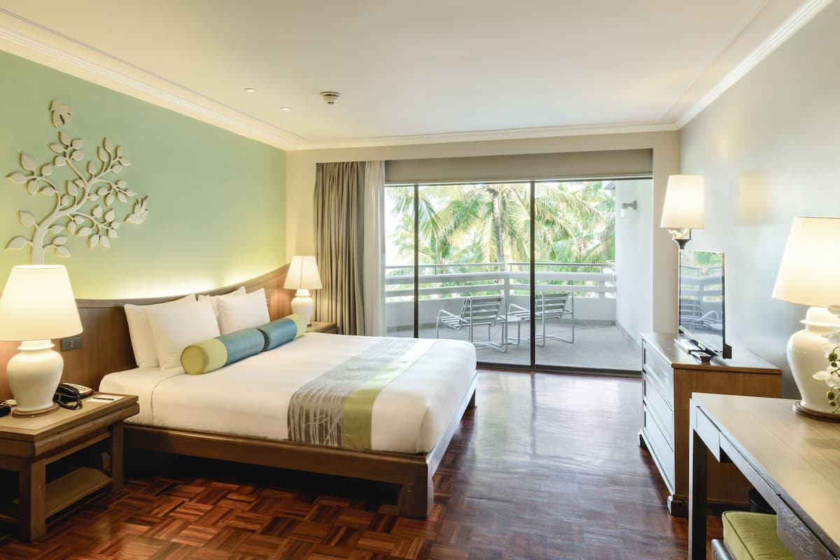 A hotel room with a large bed, a wooden nightstand, a desk with a lamp, and a balcony with a view of palm trees.