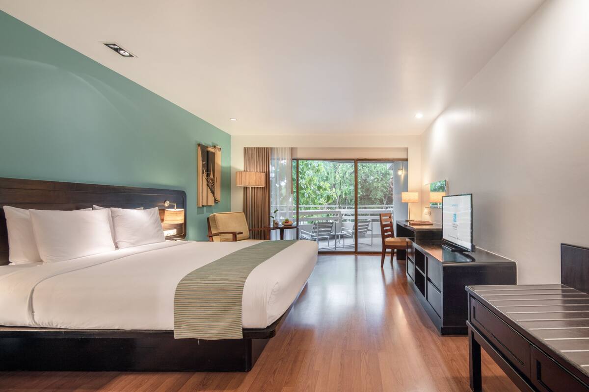 A hotel room with a large bed, a desk with a flat-screen TV, a balcony with outdoor seating, and a view of greenery.