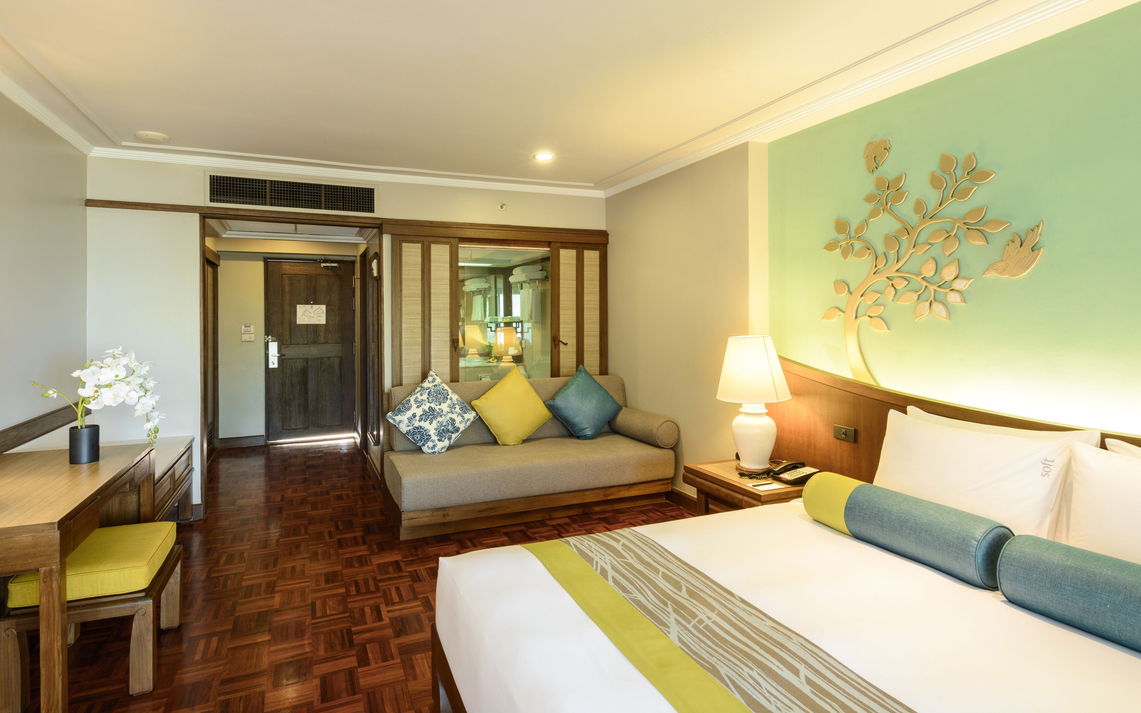 A hotel room with a bed, a desk, a chair, a sofa, a lamp, and a decorative wall piece.