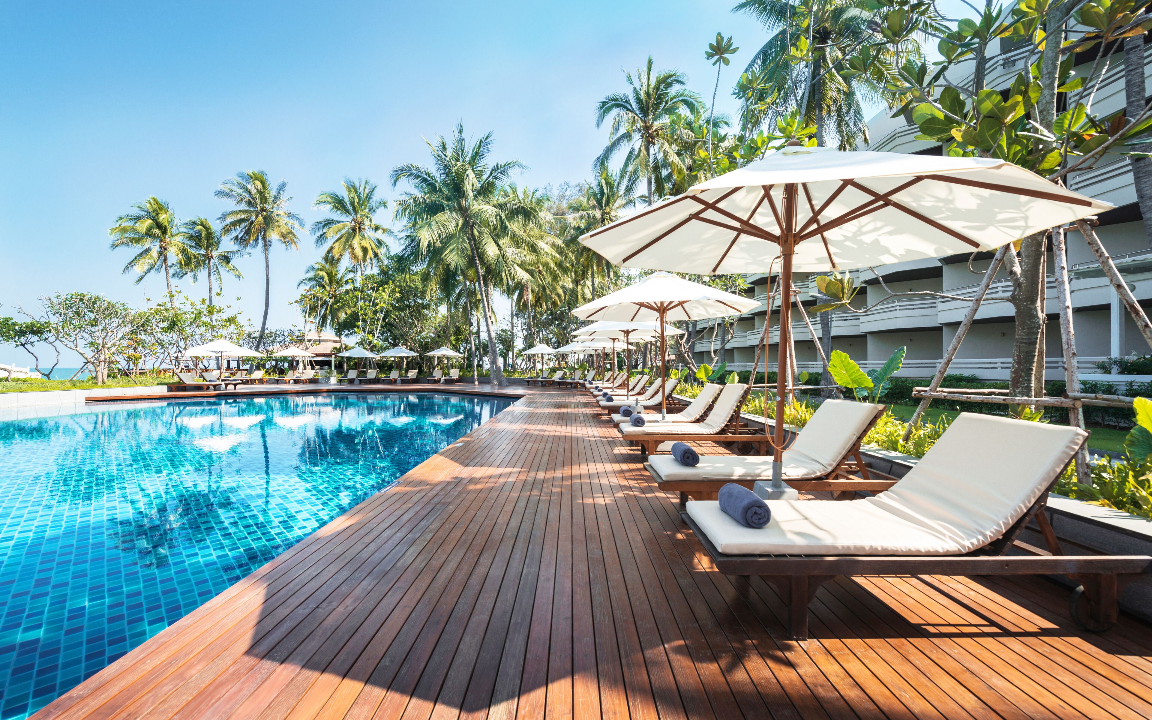 A poolside area with lounge chairs and umbrellas, surrounded by palm trees and a building in the background.