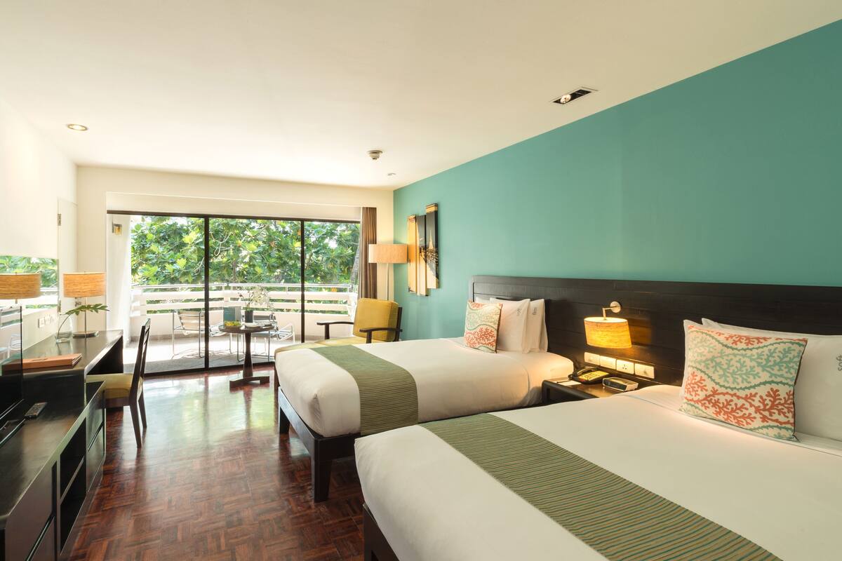 A hotel room with two beds, a desk, a chair, a lamp, and a view of greenery outside.