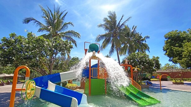 Un área de juegos acuáticos con toboganes y chorros de agua, rodeada de palmeras y cielos despejados.