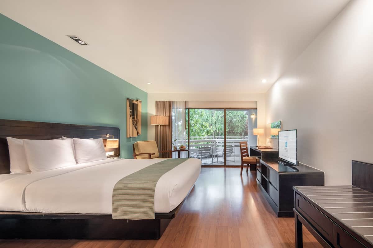 A hotel room with a large bed, a desk with a computer, a TV, and a balcony with outdoor seating.