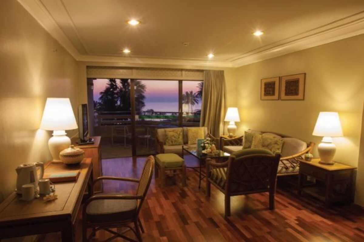 Habitación de hotel con piso de madera, un sofá, una silla, una mesita y un televisor. Hay dos lámparas sobre las mesitas de noche.