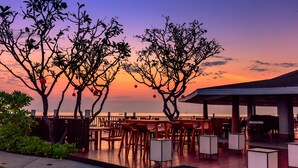 Un área de comedor al aire libre con mesas y sillas, una terraza de madera y árboles con adornos colgantes bajo un cielo de atardecer.
