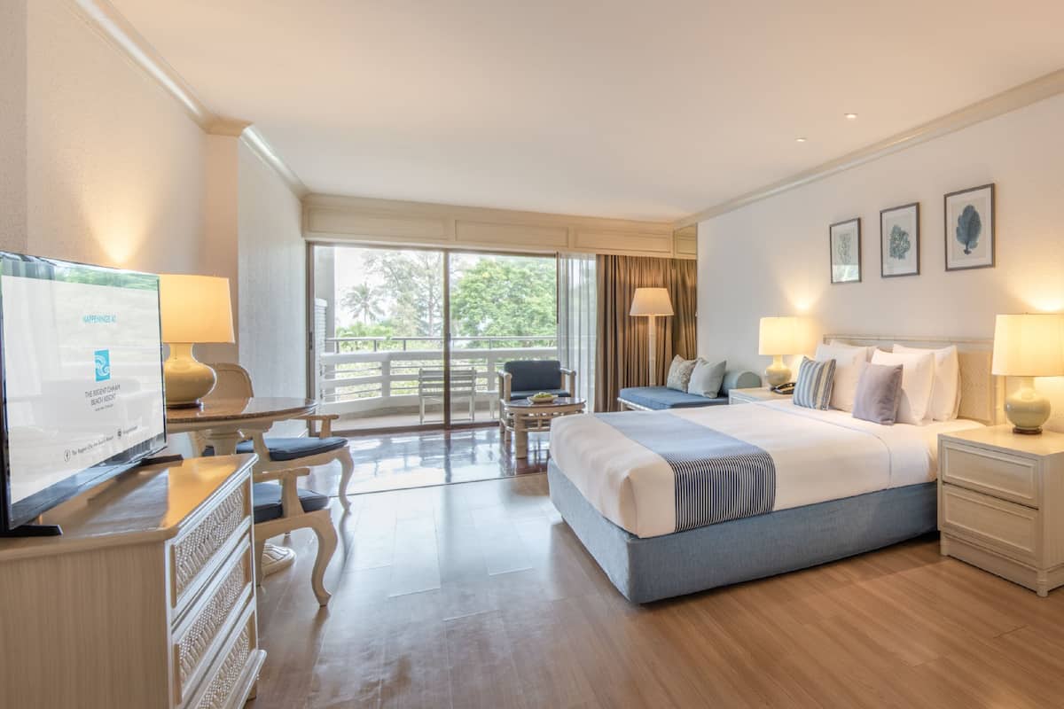A hotel room with a large bed, a desk, a chair, and a television. There is a balcony with a view of trees.