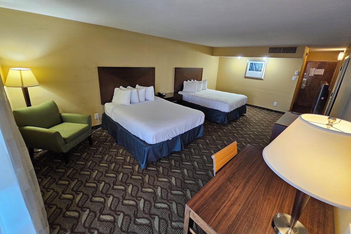 A hotel room with two beds, a green armchair, a wooden desk, and a lamp.
