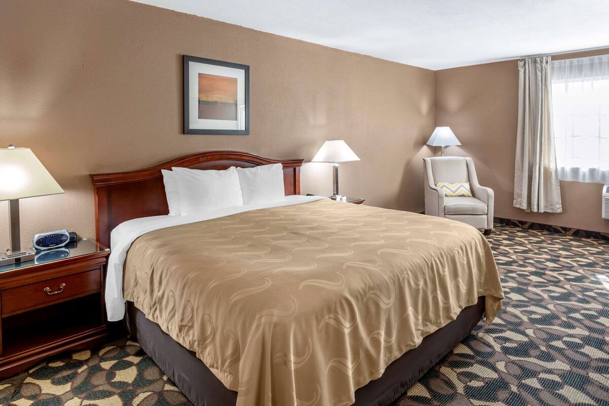 Habitación de hotel con una cama grande, dos lámparas de noche, una silla y un cuadro enmarcado en la pared.
