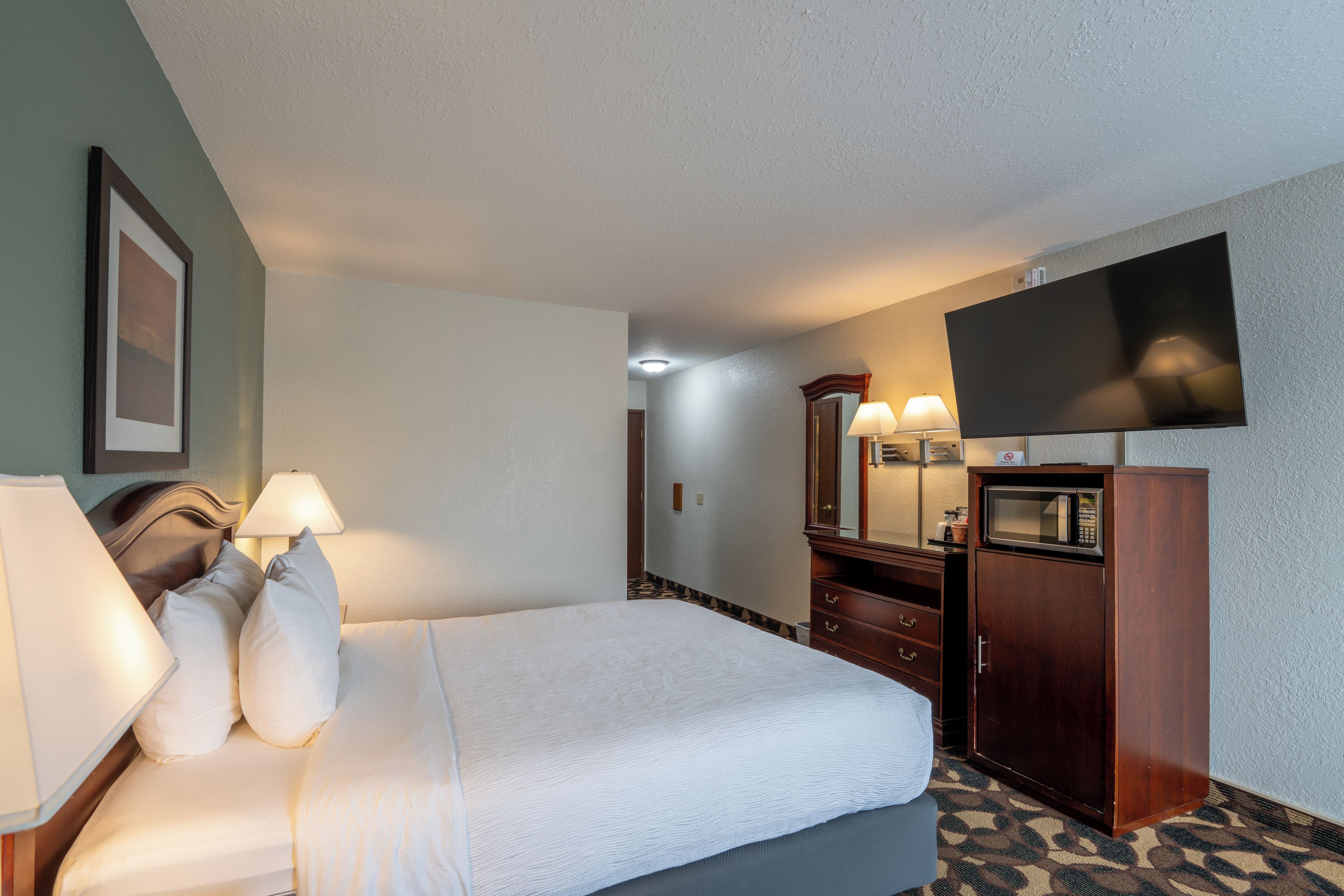 A hotel room with a bed, bedside lamps, a television, a mirror, a dresser, and a microwave.