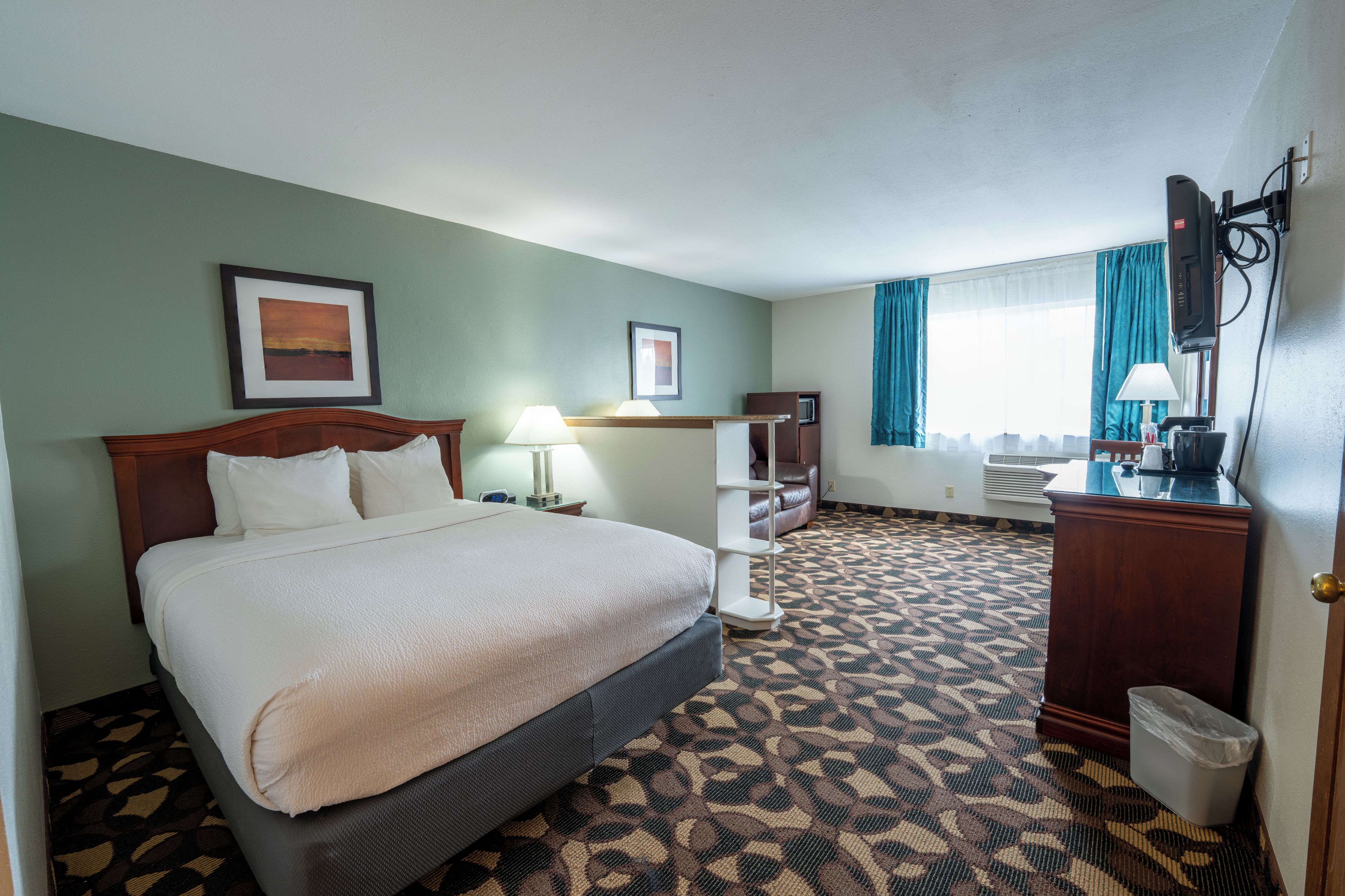 A hotel room with a bed, a desk, a chair, a waste bin, and a window with curtains.