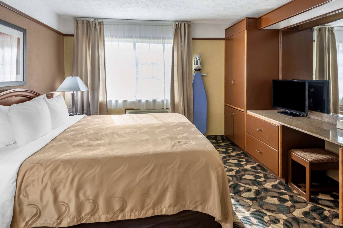 Habitación de hotel con una cama grande, un escritorio con televisor, un espejo y una ventana con cortinas.